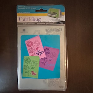 May include: A Cuttlebug cut & emboss machine kit with a clear plastic package. The kit includes a blue base with three colorful cards: pink, green, and purple, each with a floral design and text. The package also includes a small green die-cutting machine.