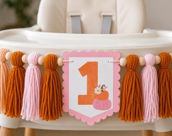 Op de afbeelding: Een kinderstoelbanner met een roze kartonnen bordje met een geschulpte rand, een grote oranje nummer één, een kleine oranje pompoen, een roze pompoen en witte bloemen. Het bordje wordt geflankeerd door afwisselende oranje en roze wollen kwastjes.