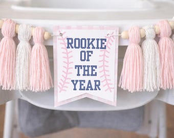 May include: A banner with pink and white tassels and wooden beads is strung across a high chair. The banner has a pennant in the center with the words "ROOKIE OF THE YEAR" in navy blue lettering, with baseball stitching accents.