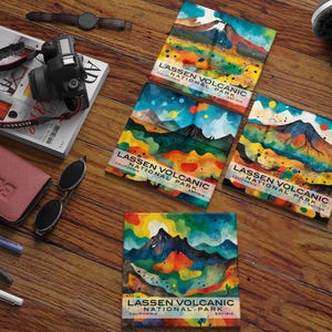 May include: A collection of colorful square art prints featuring a watercolor painting of Lassen Volcanic National Park in California. The prints display a mountain landscape with vibrant hues of blue, green, orange, and yellow. Other items include a camera, magazine, and sunglasses.