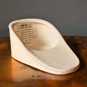 May include: An off-white ceramic slipper bed pan with a sloped design. The interior features black printed text that reads: THE NEW SLIPPER BED PAN, Slipper should be passed under patient in front between the legs, flannel cap is made for the, fastened by strings under, considerable comfort, be afforded, BRITISH IRONSTONE.