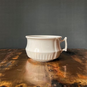 May include: White ceramic chamber pot with a single handle. The pot has a fluted design and is sitting on a wooden surface.