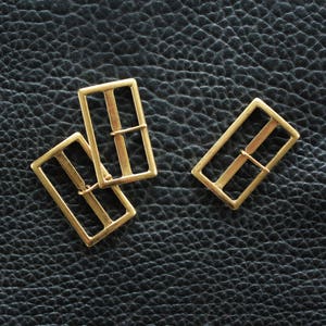 Puede incluir: Tres deslizadores de hebilla de cinturón de metal dorado. Las hebillas son rectangulares con un acabado liso y pulido.