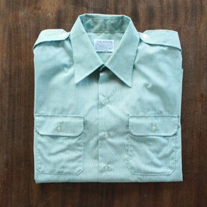 Puede incluir: Una camisa de manga larga azul claro con cuello abotonado y dos bolsillos de solapa. La camisa tiene un sutil patrón a rayas y una etiqueta blanca con el texto "Van Heusen" en el centro.