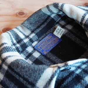 Puede incluir: Una camisa de franela a cuadros en blanco y negro con una etiqueta azul que dice "WARRANTED TO BE A PENDLETON PORTLAND OREGON PENDLETON WOOLEN MILLS 100% VIRGIN WOOL MADE IN USA" y una etiqueta blanca que dice "dry clean only".