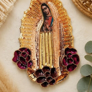 May include: An oval-shaped embroidered patch featuring a central image of the Virgin of Guadalupe with hands clasped in prayer. The design is heavily embellished with shimmering gold sequins and accented by deep purple sequined floral motifs.