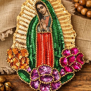 May include: A decorative religious icon featuring a depiction of the Virgin Mary. The image is adorned with gold sequins, green and red sequins, and purple and orange floral accents. The icon is set against a wooden background.
