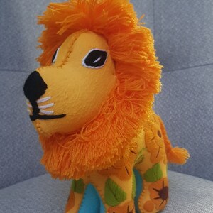 Puede incluir: Un juguete de peluche de le&oacute;n con una melena naranja brillante y un cuerpo amarillo. El le&oacute;n lleva un atuendo azul y verde con girasoles bordados.