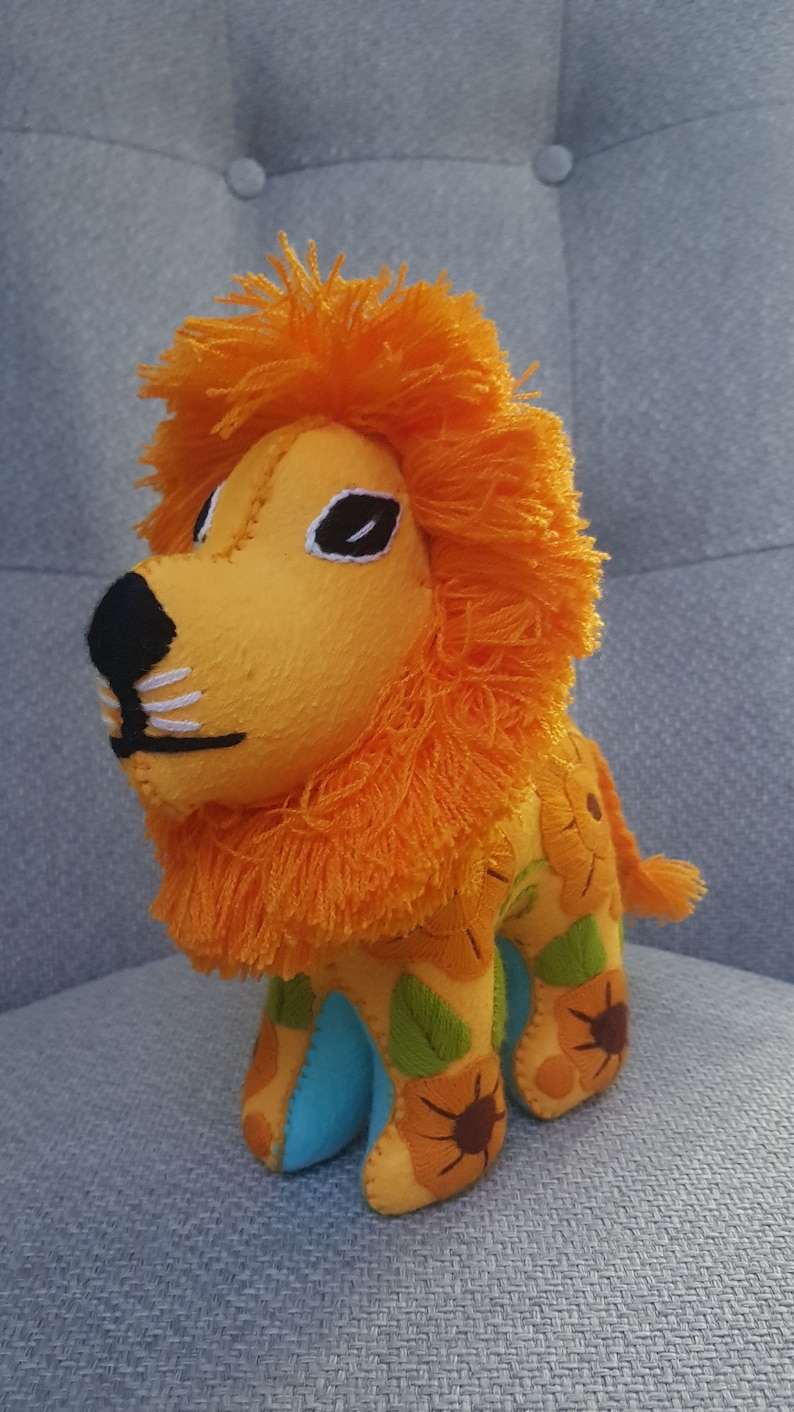 Puede incluir: Un juguete de peluche de le&oacute;n con una melena naranja brillante y un cuerpo amarillo. El le&oacute;n lleva un atuendo azul y verde con girasoles bordados.