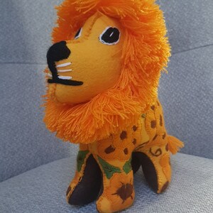Puede incluir: Un juguete de peluche de le&oacute;n naranja con nariz marr&oacute;n y ojos negros. El le&oacute;n est&aacute; bordado con un patr&oacute;n floral y tiene una melena naranja esponjosa.