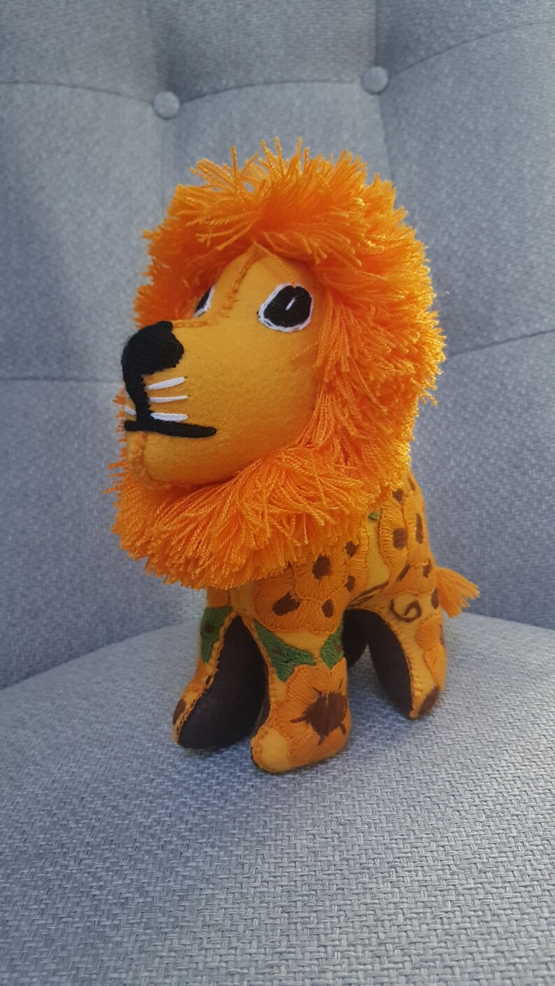 Puede incluir: Un juguete de peluche de le&oacute;n naranja con nariz marr&oacute;n y ojos negros. El le&oacute;n est&aacute; bordado con un patr&oacute;n floral y tiene una melena naranja esponjosa.