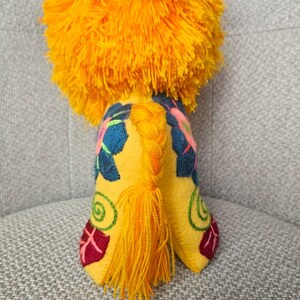May include: A plush lion toy featuring a vibrant orange yarn mane and a yellow felt body. The body is decorated with embroidered floral patterns in dark blue, pink, green, and red. A braided yellow yarn tail hangs down the center back.