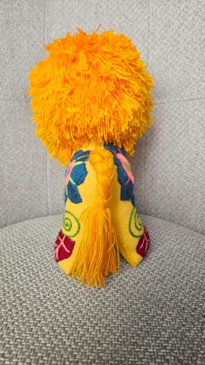 May include: A plush lion toy featuring a vibrant orange yarn mane and a yellow felt body. The body is decorated with embroidered floral patterns in dark blue, pink, green, and red. A braided yellow yarn tail hangs down the center back.