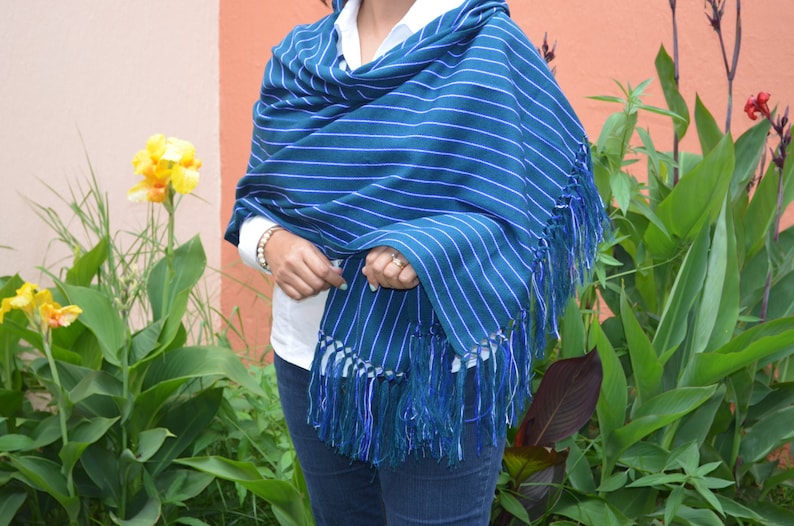 Puede incluir: Un chal a rayas azules y blancas con flecos. El chal est&aacute; drapeado sobre una persona que lleva una camisa blanca y vaqueros azules. El fondo incluye plantas y flores verdes.