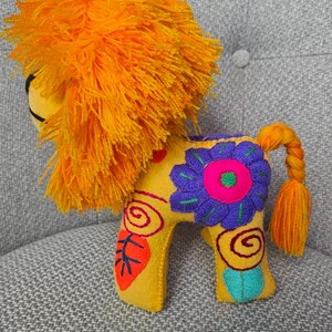 May include: A yellow felt lion plush toy featuring a vibrant orange yarn mane and a braided tail. The body is decorated with colorful embroidered floral and spiral patterns in shades of purple, pink, red, and teal, showcasing detailed stitching.