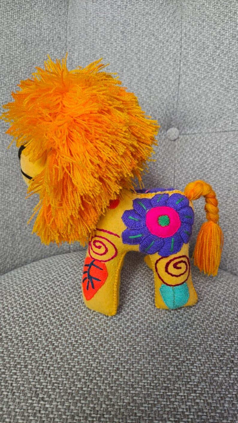 May include: A yellow felt lion plush toy featuring a vibrant orange yarn mane and a braided tail. The body is decorated with colorful embroidered floral and spiral patterns in shades of purple, pink, red, and teal, showcasing detailed stitching.