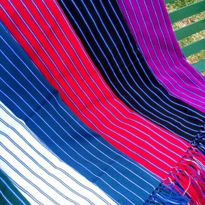Puede incluir: Una colecci&oacute;n de coloridas mantas tejidas a rayas, exhibidas en un banco verde. Las mantas presentan varios colores, incluyendo rojo, azul, negro, blanco y morado, con rayas blancas. Cada manta tiene un borde con flecos.