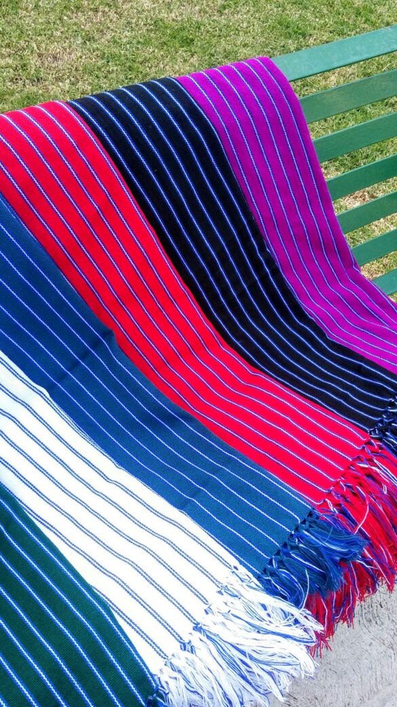 Puede incluir: Una colecci&oacute;n de coloridas mantas tejidas a rayas, exhibidas en un banco verde. Las mantas presentan varios colores, incluyendo rojo, azul, negro, blanco y morado, con rayas blancas. Cada manta tiene un borde con flecos.