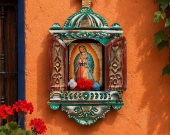 May include: A wooden wall-mounted shrine featuring a central image of the Virgin of Guadalupe. The frame is painted in distressed green and red with a gold cross on top. Small fabric flowers sit at the base of the central religious icon.