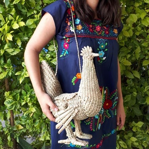 May include: A woven straw handbag shaped like a rooster. The bag has a long strap and is being held by a person wearing a blue embroidered dress.