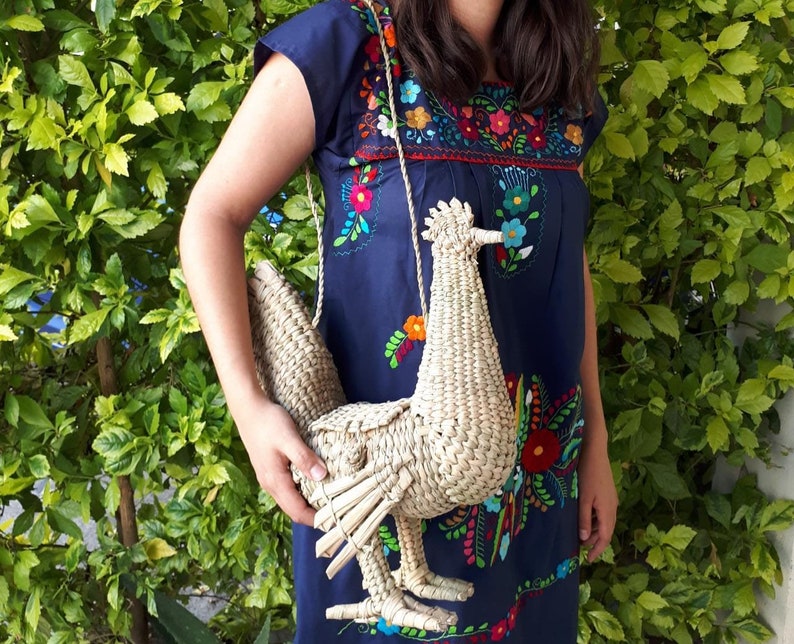 May include: A woven straw handbag shaped like a rooster. The bag has a long strap and is being held by a person wearing a blue embroidered dress.