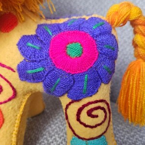 May include: A yellow felt animal figurine featuring a vibrant purple and hot pink embroidered flower on its side. The creature has a bright orange yarn mane, a braided orange and yellow tail, and additional red and teal spiral embroidery.