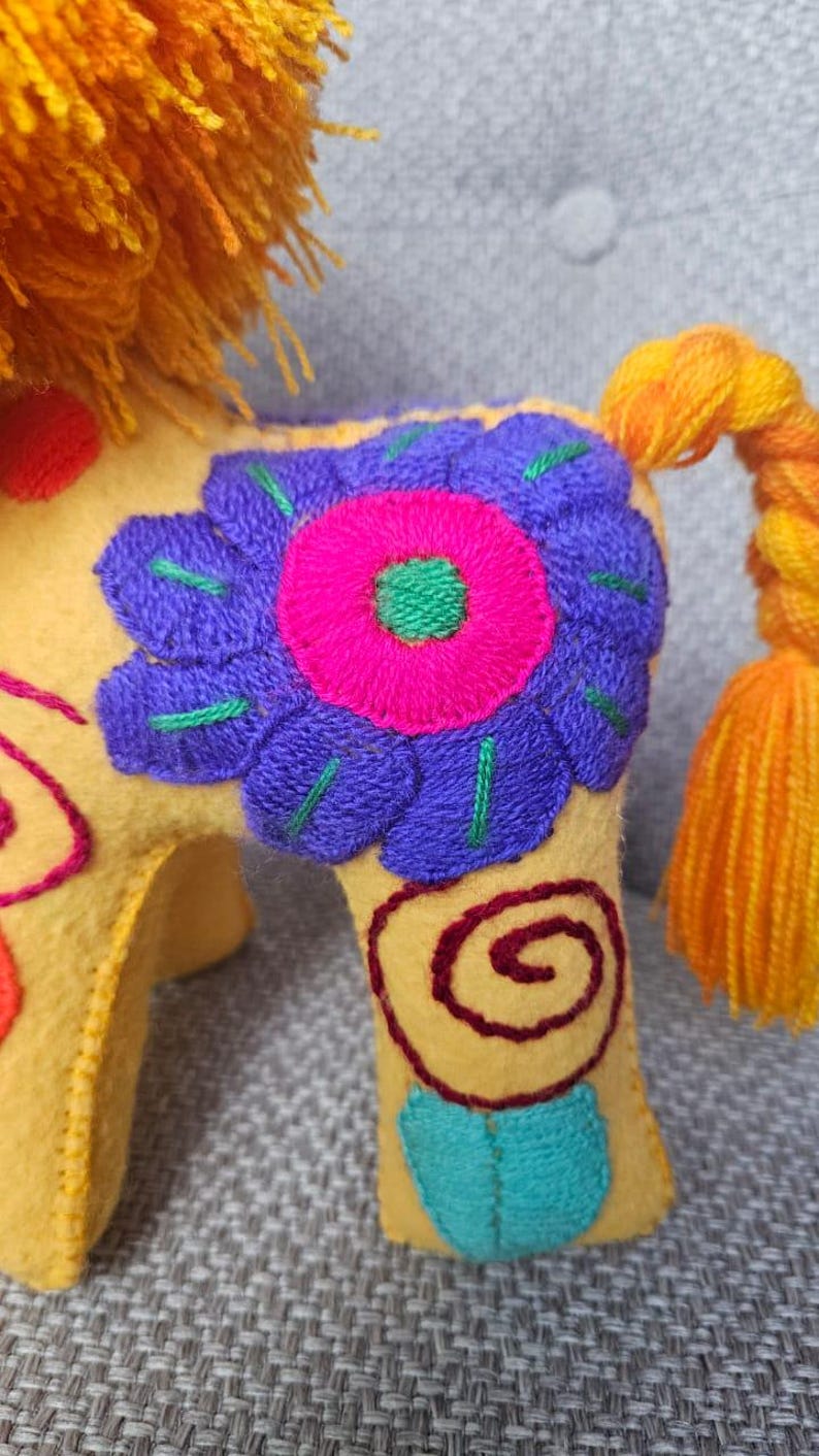 May include: A yellow felt animal figurine featuring a vibrant purple and hot pink embroidered flower on its side. The creature has a bright orange yarn mane, a braided orange and yellow tail, and additional red and teal spiral embroidery.