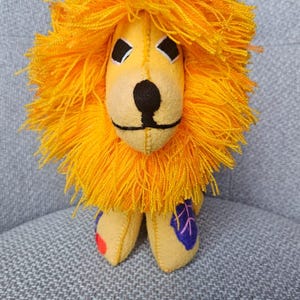 May include: A stuffed lion toy made of yellow fabric with a prominent, thick, bright orange yarn mane. The face features black embroidered eyes, a black nose, and a stitched mouth. One paw has a red circular patch, and another has a purple leaf shape.