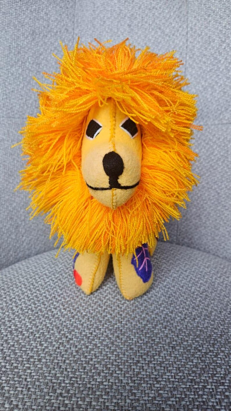 May include: A stuffed lion toy made of yellow fabric with a prominent, thick, bright orange yarn mane. The face features black embroidered eyes, a black nose, and a stitched mouth. One paw has a red circular patch, and another has a purple leaf shape.
