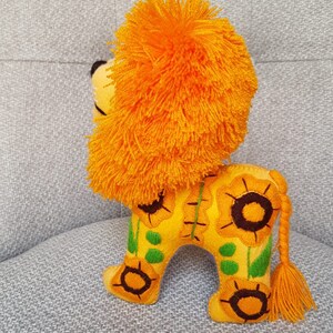 Puede incluir: Un juguete de peluche de le&oacute;n con una melena de color naranja brillante y un cuerpo amarillo. El cuerpo est&aacute; decorado con girasoles bordados y hojas verdes.