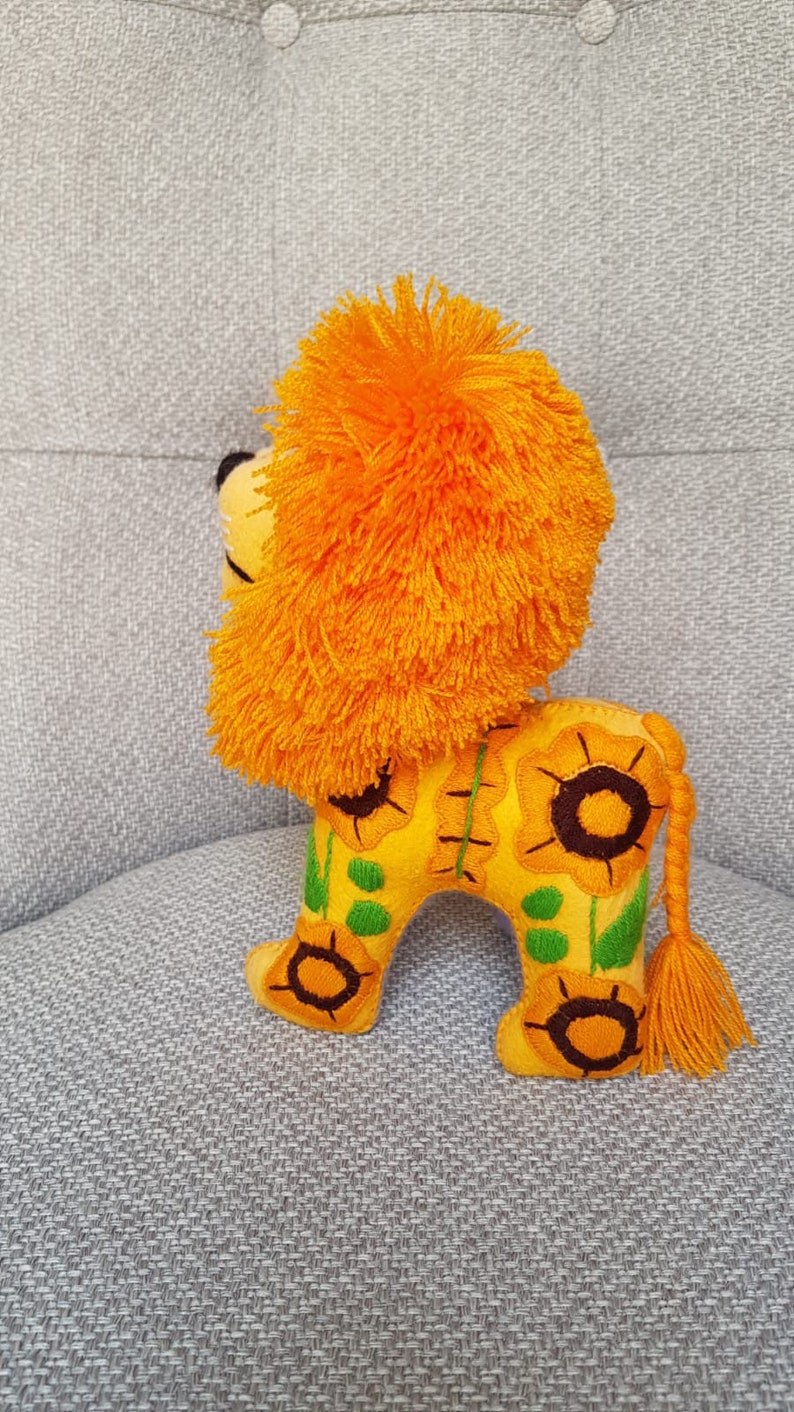 Puede incluir: Un juguete de peluche de le&oacute;n con una melena de color naranja brillante y un cuerpo amarillo. El cuerpo est&aacute; decorado con girasoles bordados y hojas verdes.