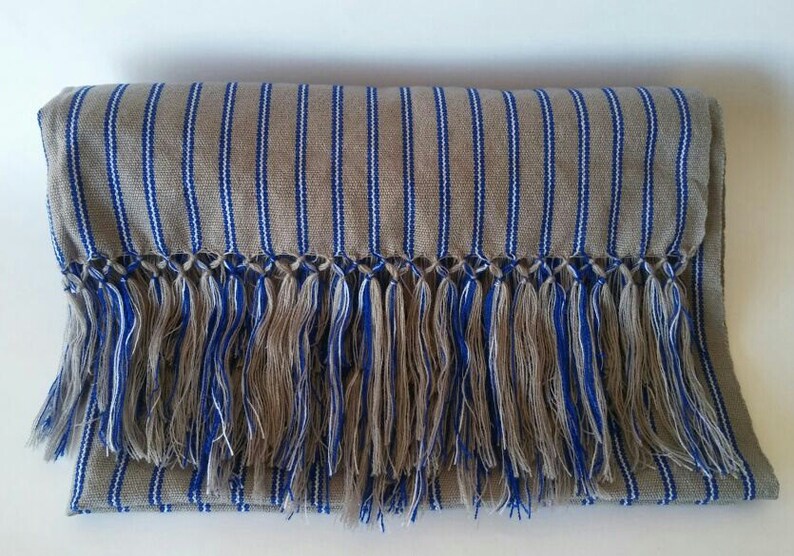 Puede incluir: Una bufanda beige doblada con rayas verticales azules y blancas. La bufanda tiene un fleco de hilos azules, blancos y beige a lo largo del borde inferior. La bufanda est&aacute; hecha de tela tejida.