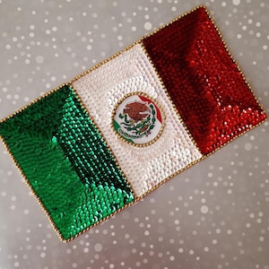 May include: A rectangular Mexican flag made of sequins. The flag features green, white, and red sections, with a gold beaded border. The center has a circular emblem with the Mexican coat of arms.