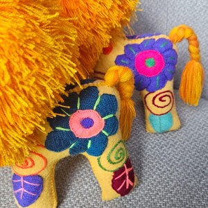 May include: Two yellow felt animal figures featuring bright orange yarn manes and tails. The bodies are decorated with embroidered floral patterns in blue, purple, pink, and green, including spiral and leaf motifs on the legs and torso.