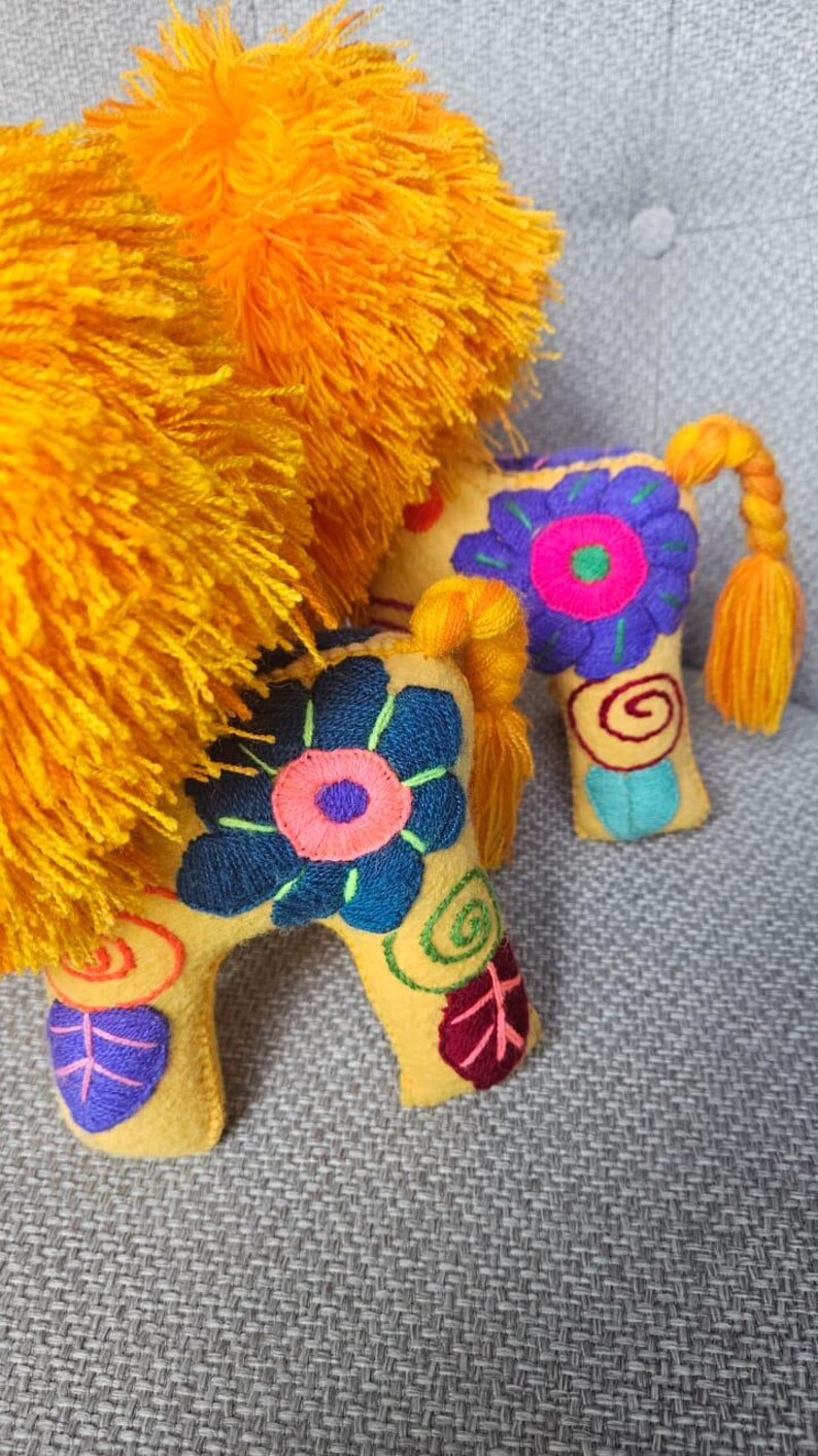 May include: Two yellow felt animal figures featuring bright orange yarn manes and tails. The bodies are decorated with embroidered floral patterns in blue, purple, pink, and green, including spiral and leaf motifs on the legs and torso.