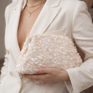 May include: A cream-colored clutch covered in small, round sequins. The clutch is held against a white blazer, with gold necklaces visible. The sequins create a textured surface, reflecting light. The overall aesthetic is elegant and suitable for special occasions.
