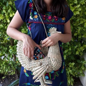 May include: A woven straw handbag shaped like a rooster, with a blue and floral embroidered dress in the background.