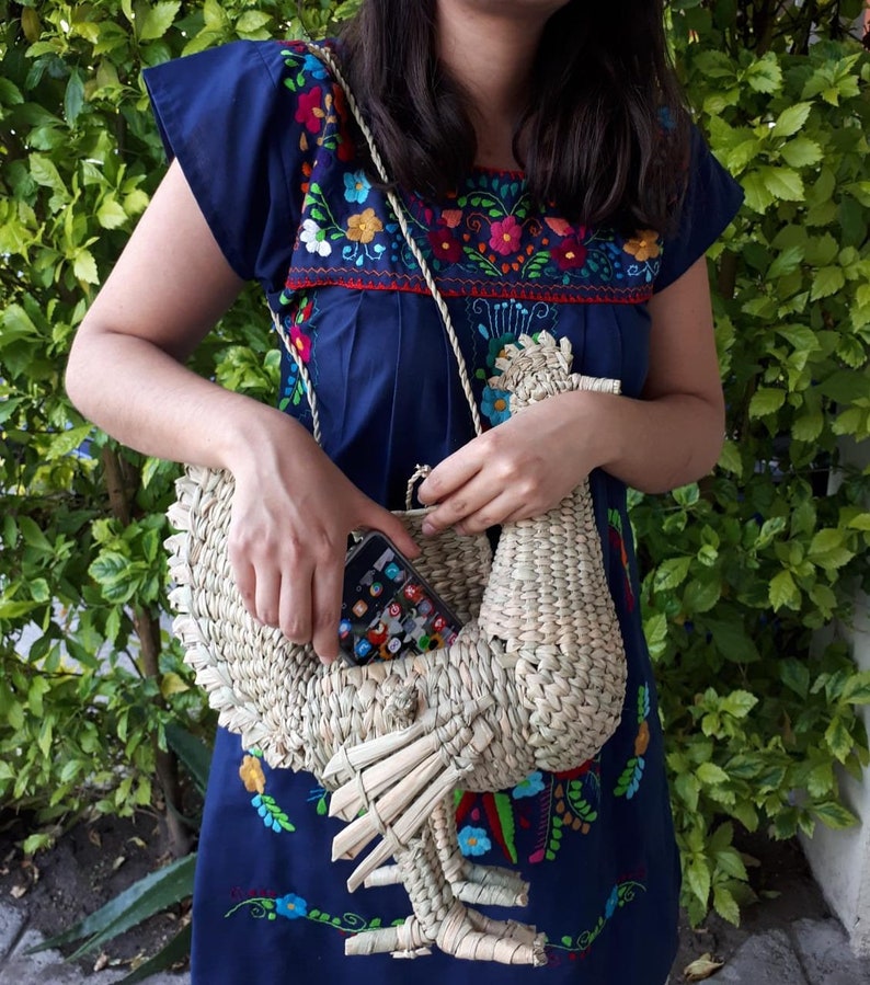 May include: A woven straw handbag shaped like a rooster, with a blue and floral embroidered dress in the background.