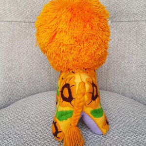 Puede incluir: Un le&oacute;n de peluche con una melena de color naranja brillante y un cuerpo estampado en amarillo, verde y marr&oacute;n. El le&oacute;n tiene una cola larga trenzada con un borla.