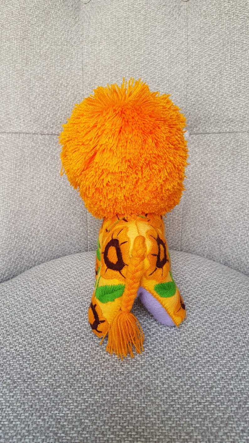 Puede incluir: Un le&oacute;n de peluche con una melena de color naranja brillante y un cuerpo estampado en amarillo, verde y marr&oacute;n. El le&oacute;n tiene una cola larga trenzada con un borla.