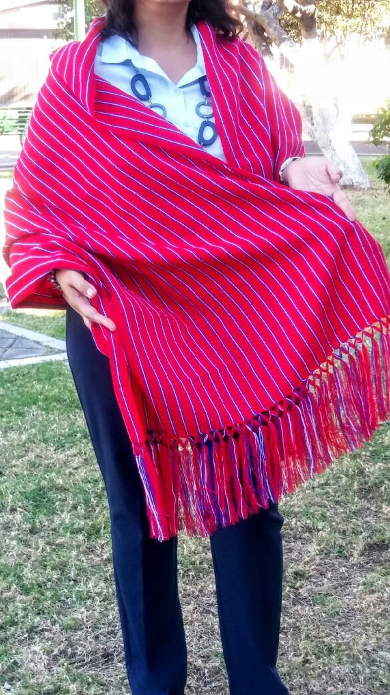 Puede incluir: Un chal rojo vibrante con rayas verticales blancas y un borde con flecos. El chal est&aacute; drapeado sobre una camisa azul claro y pantalones azul oscuro. La franja incluye hilos rojos, azules y dorados.