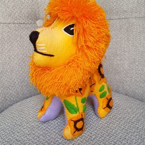 Puede incluir: Un juguete de peluche de le&oacute;n con una melena naranja brillante y un cuerpo amarillo con girasoles bordados. El le&oacute;n tiene una base de fieltro morado y est&aacute; de pie sobre una superficie gris texturizada.