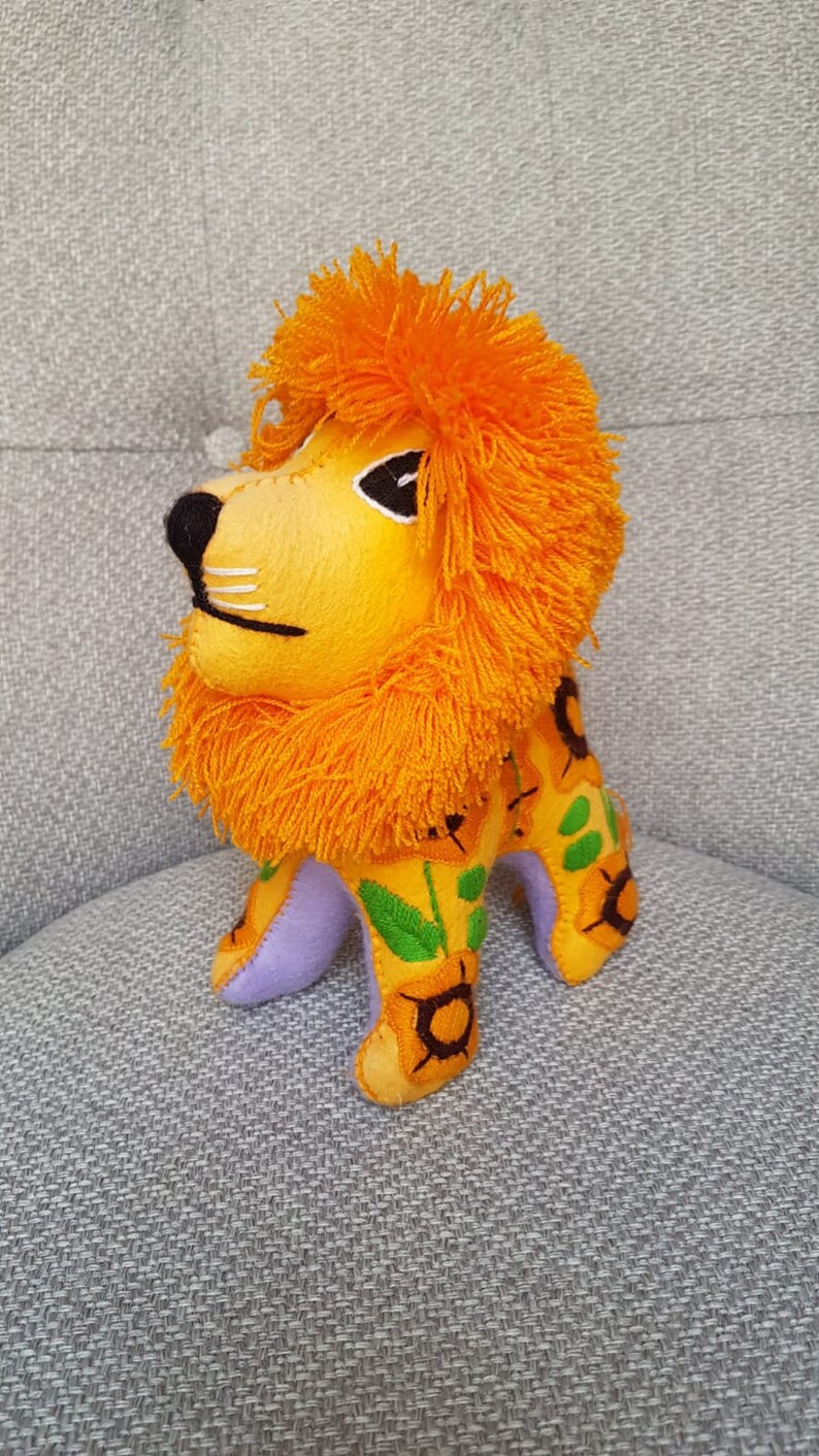 Puede incluir: Un juguete de peluche de le&oacute;n con una melena naranja brillante y un cuerpo amarillo con girasoles bordados. El le&oacute;n tiene una base de fieltro morado y est&aacute; de pie sobre una superficie gris texturizada.