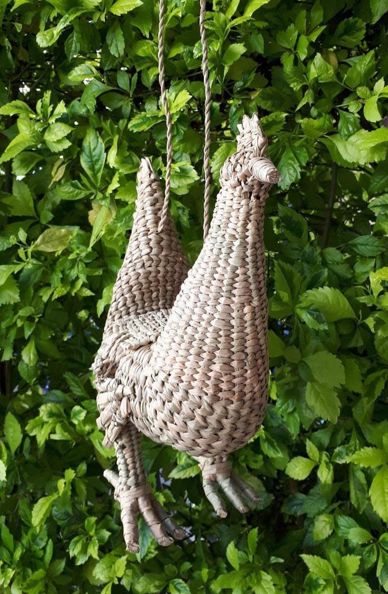 May include: A woven straw chicken-shaped hanging decoration with a brown rope. The chicken is hanging from a tree branch.