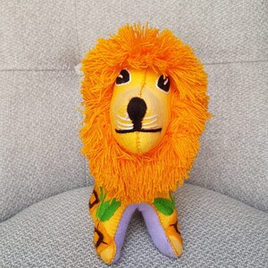Puede incluir: Un juguete de peluche de le&oacute;n naranja con la cara amarilla, la nariz negra y los bigotes blancos. El le&oacute;n est&aacute; sentado sobre una superficie de tela gris.