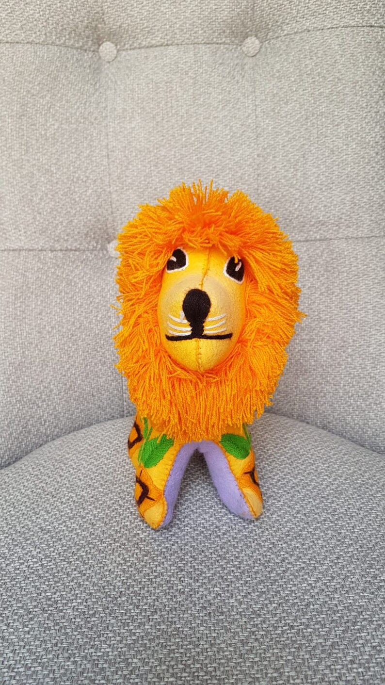 Puede incluir: Un juguete de peluche de le&oacute;n naranja con la cara amarilla, la nariz negra y los bigotes blancos. El le&oacute;n est&aacute; sentado sobre una superficie de tela gris.