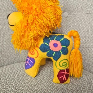May include: A yellow felt lion plush toy featuring a vibrant orange yarn mane and a braided tail with a tassel. The body is decorated with embroidered floral and spiral patterns in shades of dark blue, pink, purple, and green on its sides.