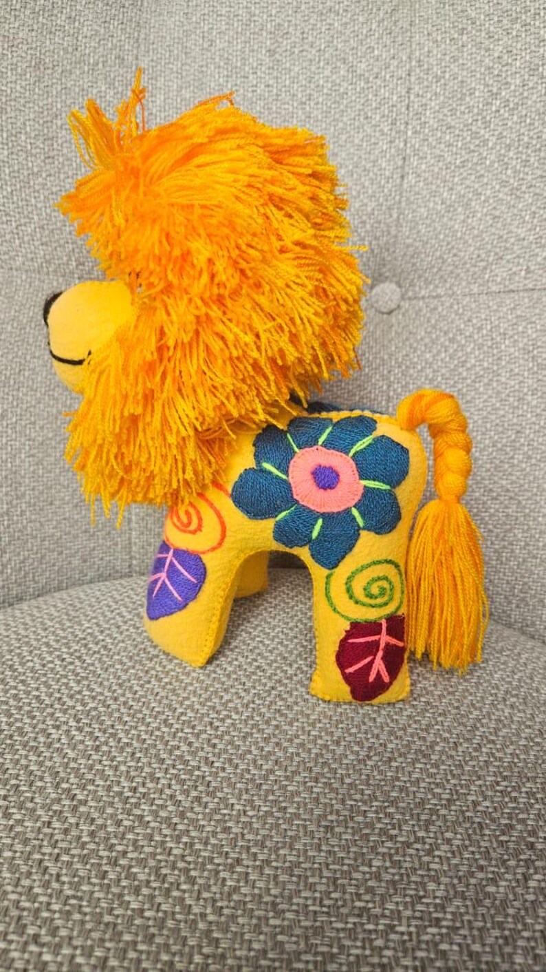 May include: A yellow felt lion plush toy featuring a vibrant orange yarn mane and a braided tail with a tassel. The body is decorated with embroidered floral and spiral patterns in shades of dark blue, pink, purple, and green on its sides.
