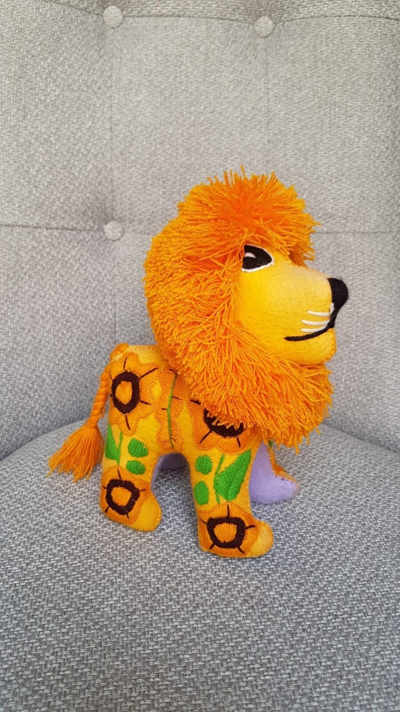 Puede incluir: Un le&oacute;n de peluche naranja con una melena esponjosa, bordado con girasoles amarillos y hojas verdes. El le&oacute;n tiene una parte inferior morada y una cola larga naranja.