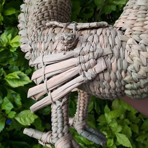 May include: A woven straw rooster figurine in shades of brown and tan. The rooster has a long tail and a detailed head with a comb and wattle. The rooster is standing on two legs with three toes each.